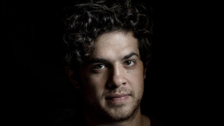 A white man with dark hair stands in front of a dark background