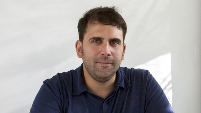 A man wears a blue polo shirt and smiles facing the camera.