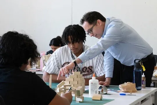 White man leaned over a desk working with a Black male high school student and his model