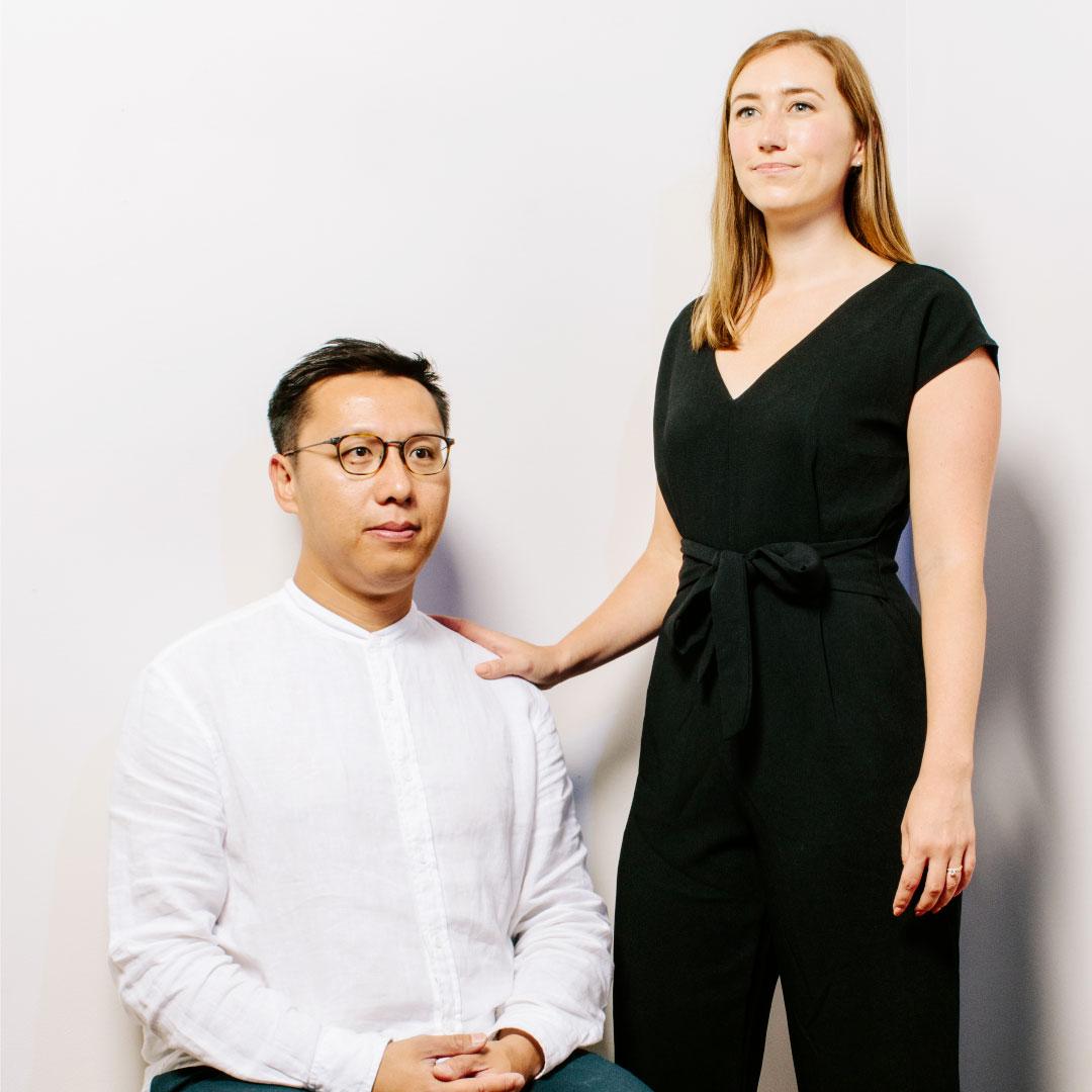 Photo of dark haired man wearing glasses sitting and woman with red hair standing next to him